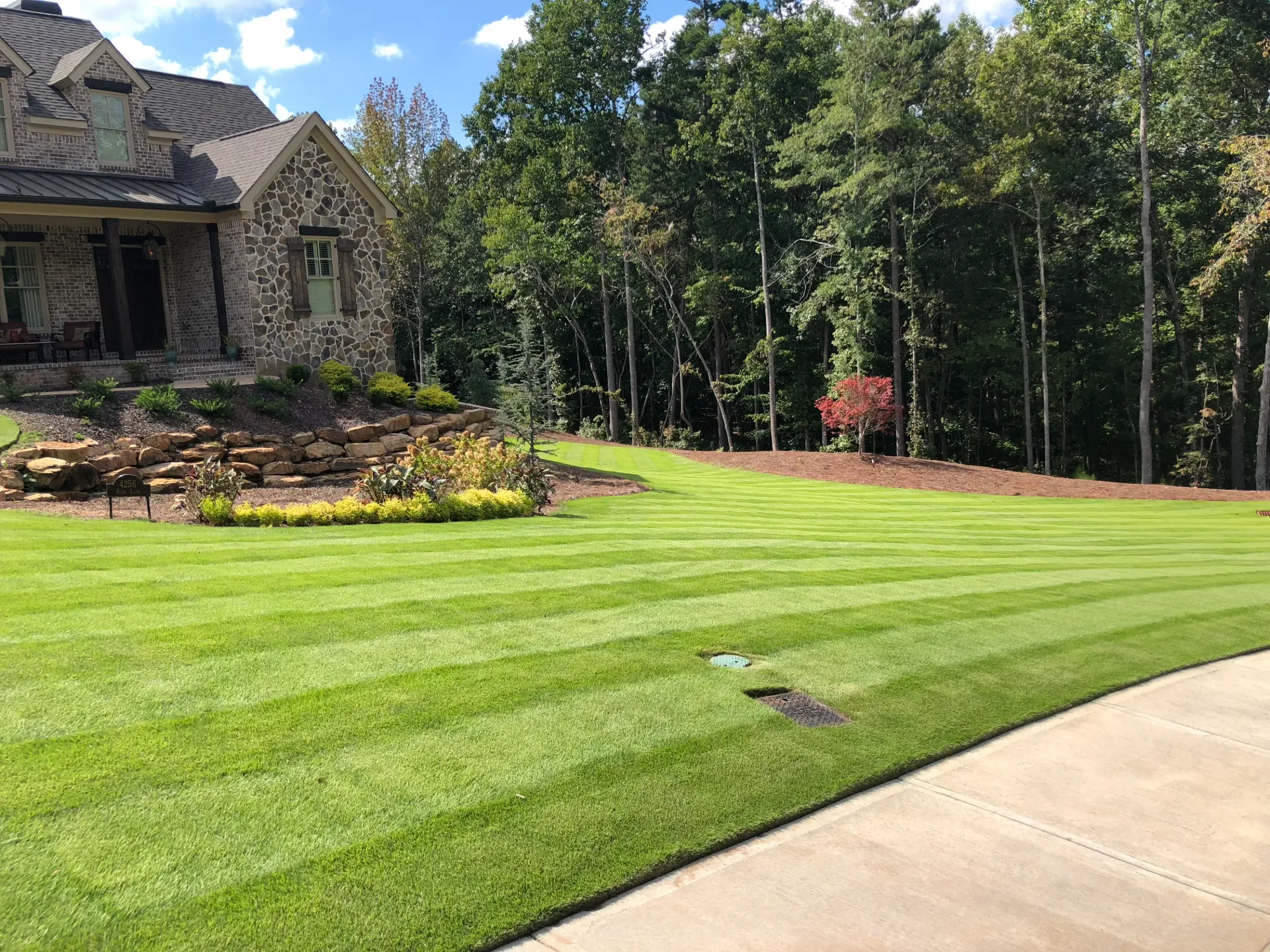A wide striped lawn wrapping around a brick and stone house at the edge of the woods