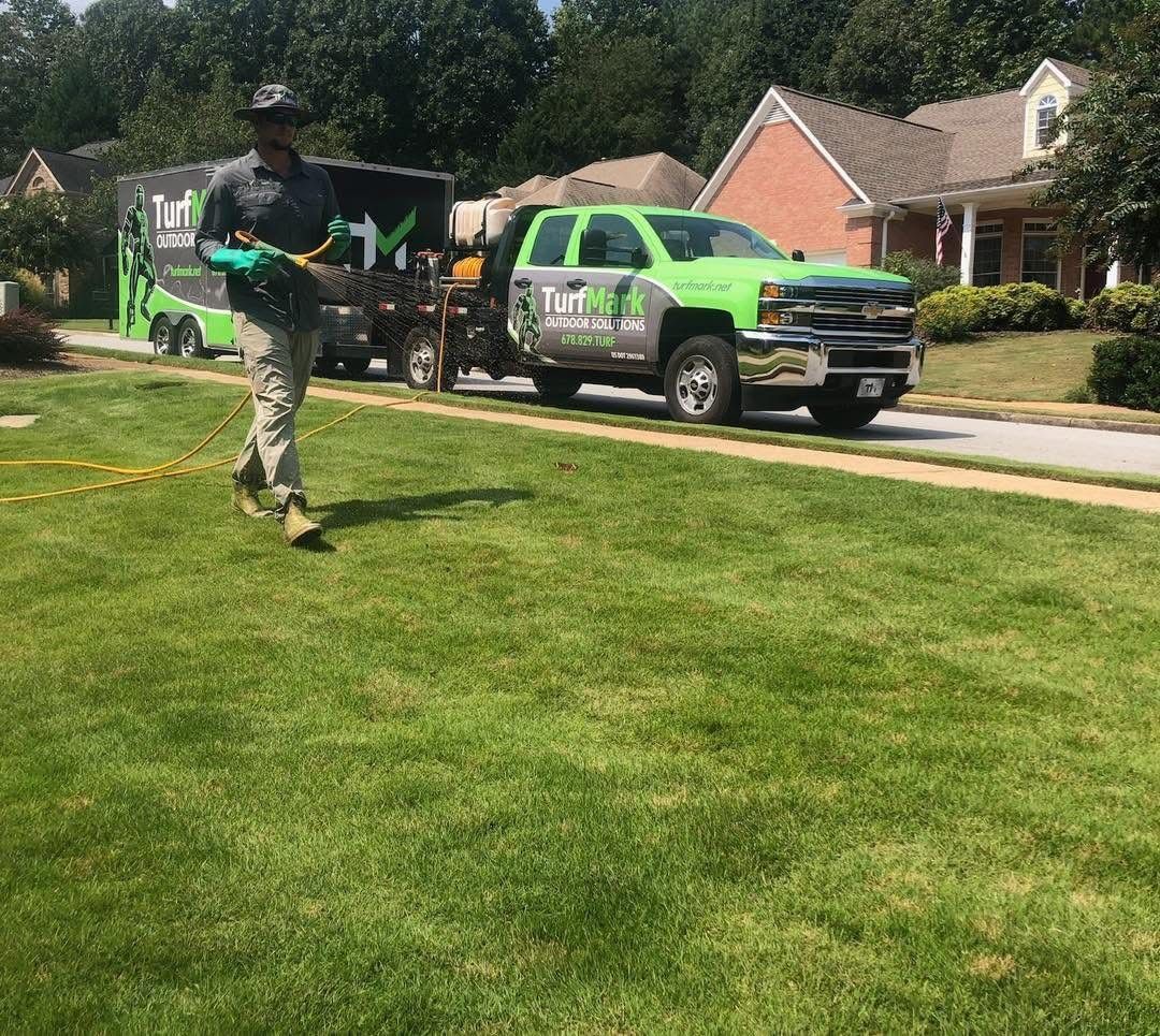 A TurfMark technician treating a front lawn, with a green TurfMark truck and trailer at the curb