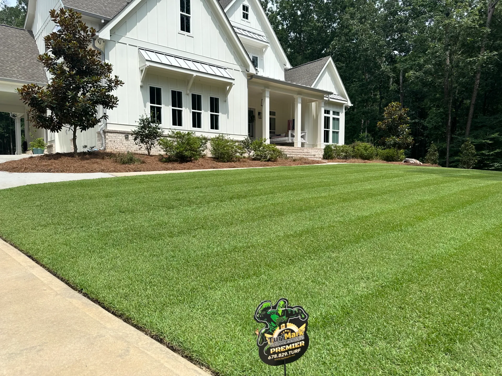 A striped lawn in front of a white farmhouse, with a TurfMark sign in the grass